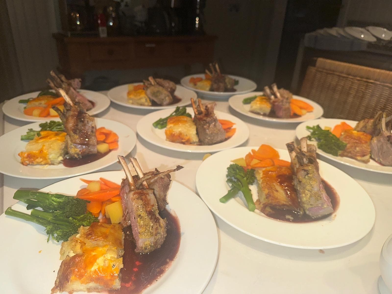 Rows of plated herb-crusted rack of lamb standing on dauphinoise potato with broccolini, carrots and a red-wine jus, laid out for service on a marble table.